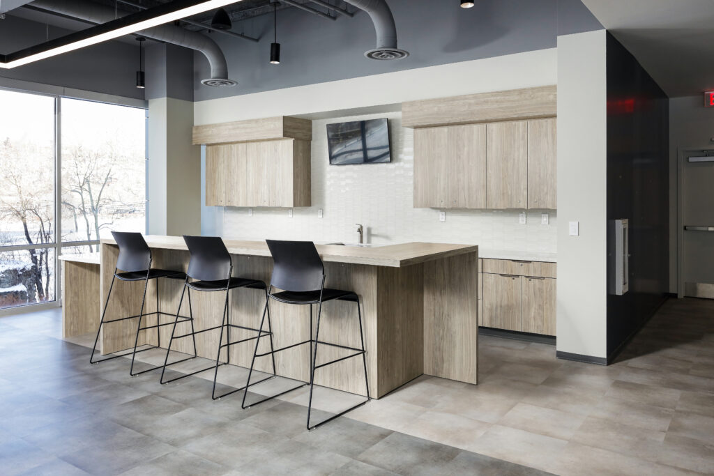Kitchenette with light wood cabinets, a long counter, and three black bar stools.