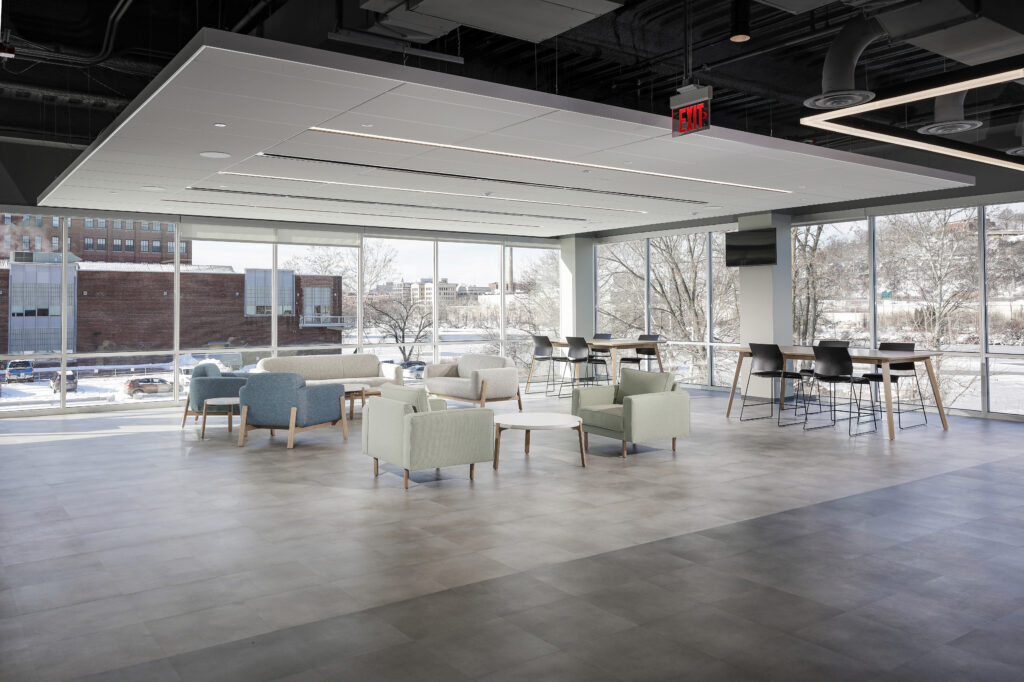 Open-plan office lounge area with modern armchairs, a sofa, small coffee tables, and high-top tables by floor-to-ceiling windows.