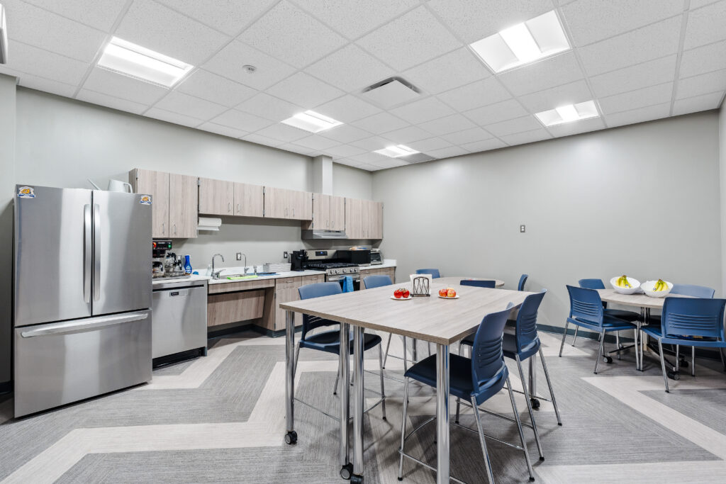 An office breakroom with a stainless steel refrigerator, kitchen counter, and two tables with blue chairs.