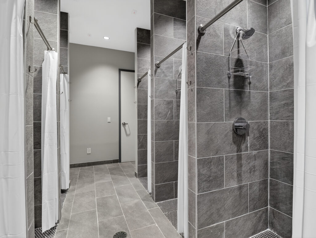 A row of shower stalls featuring gray tiled walls, white curtains, and chrome shower fixtures.