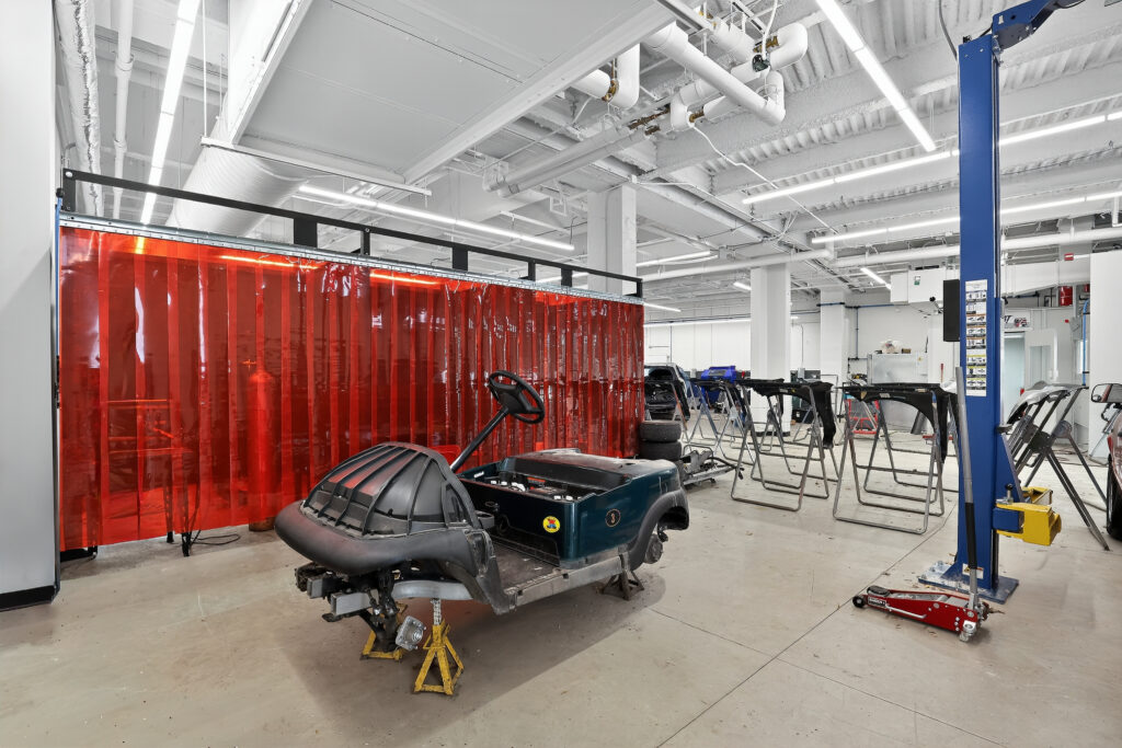 A disassembled golf cart chassis supported by jack stands in an auto body shop with red plastic privacy curtains.