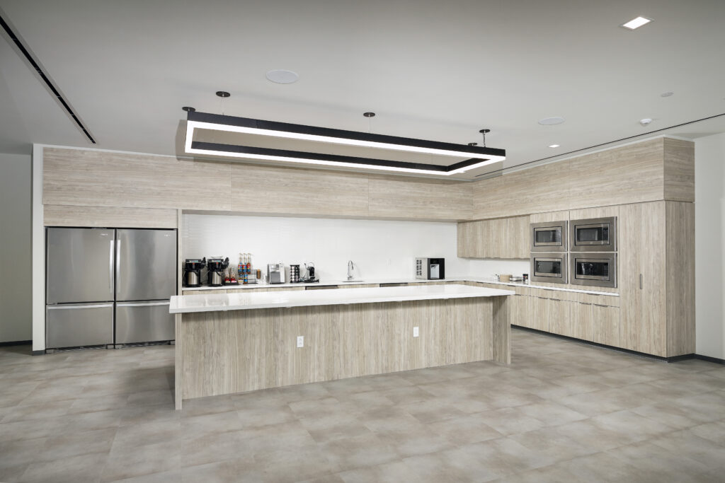 Modern office kitchen featuring a large central island, light wood cabinetry, stainless steel appliances, and a rectangular pendant light fixture.