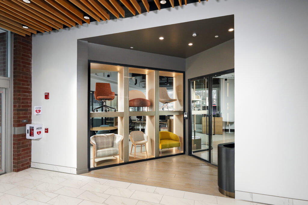 A glass-enclosed display alcove featuring six modern accent chairs, next to an AED cabinet in a building lobby.