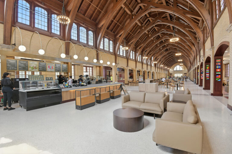 Interior of a large train station terminal with a vaulted wood ceiling, featuring a coffee shop service counter and a lounge area with beige sofas.