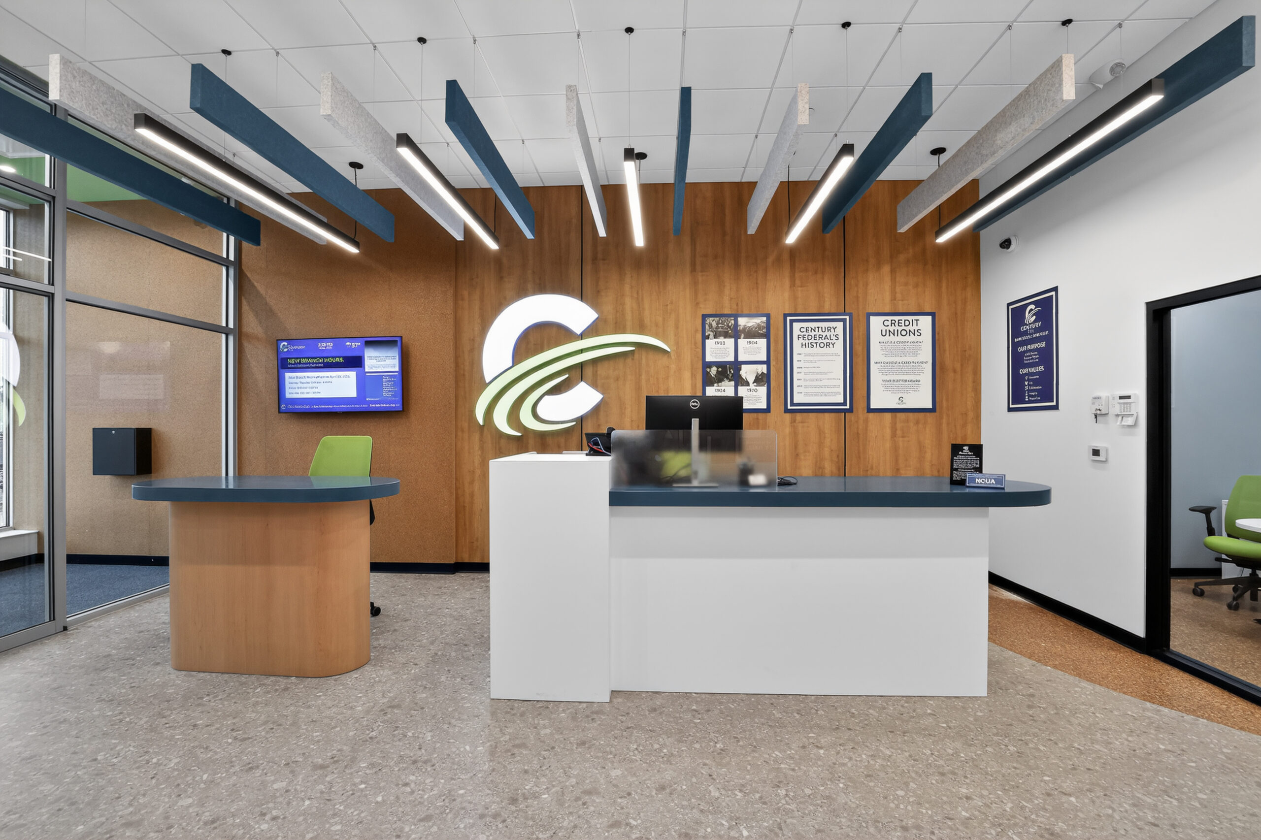 Reception area of a credit union with a white desk, wood-paneled wall featuring a large logo, informational signage, and ceiling-mounted sound baffles.