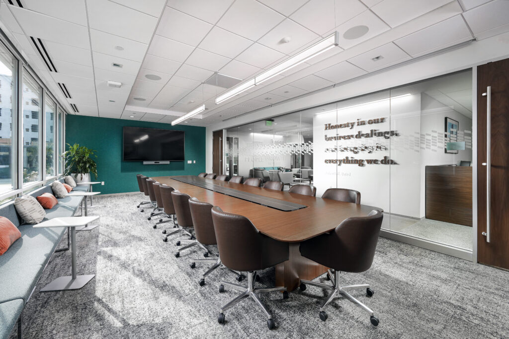 A conference room featuring a long wooden table surrounded by brown swivel chairs, with a television on a teal wall and a glass partition displaying an inspirational quote.