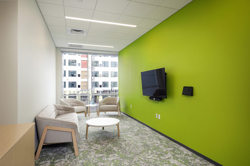 Small meeting room with a sofa, two chairs, a coffee table, and a wall-mounted television against a bright green wall.