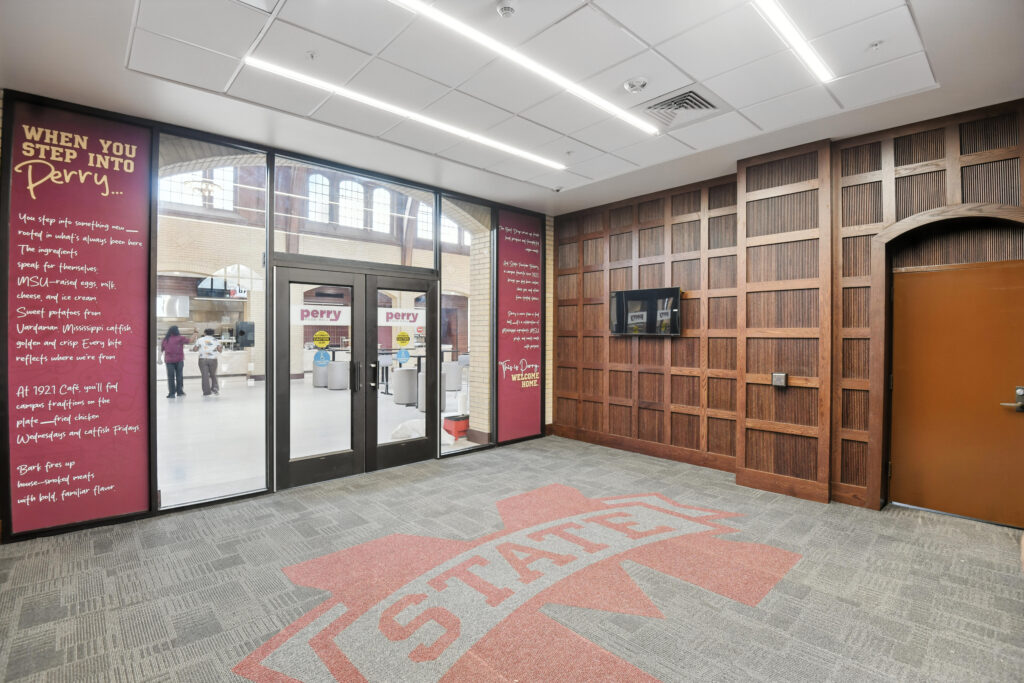 Entrance to Perry Cafeteria with a large State logo on the carpet, wood-paneled walls, and informational text on the glass.