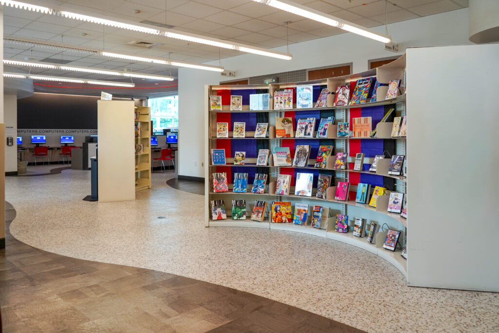 A curved library bookshelf filled with manga and comic books in a public library, with a computer station in the background.