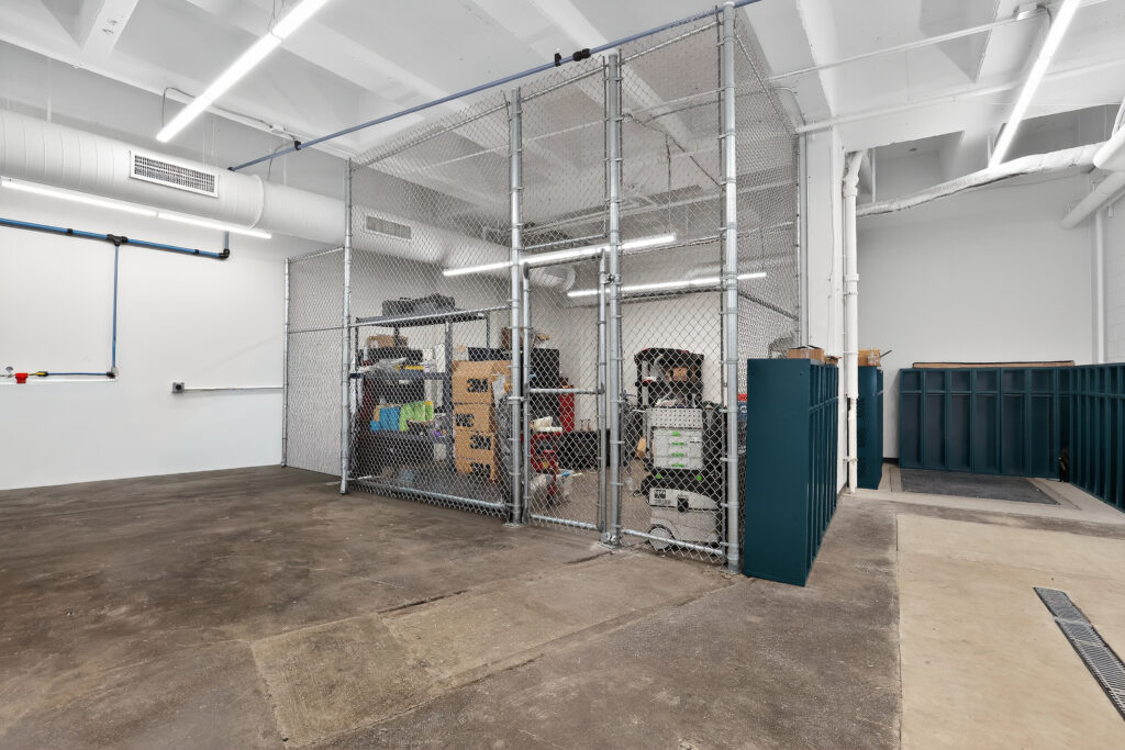 A secure chain-link storage cage inside an industrial workspace with concrete floors and open-ceiling ducts.