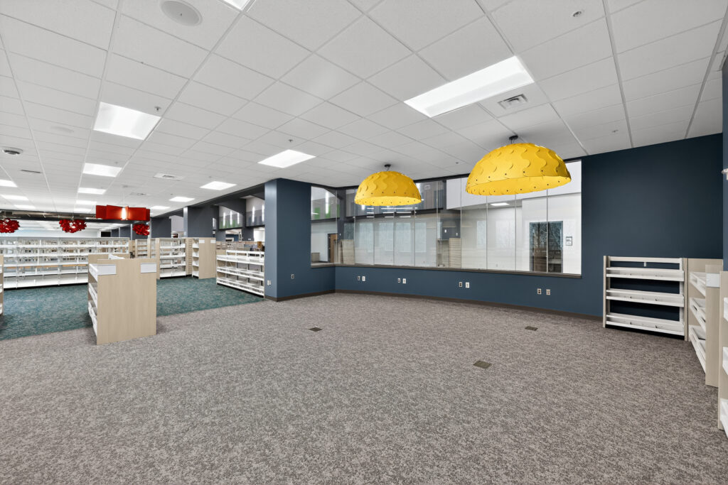Open area of a library with gray carpet, empty bookshelves, and two large, yellow dome-shaped pendant lights hanging in front of a glass-partitioned office.