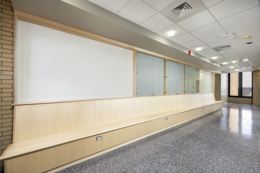 A long wooden bench built against a wall featuring a large white board and glass display cases in a hallway.