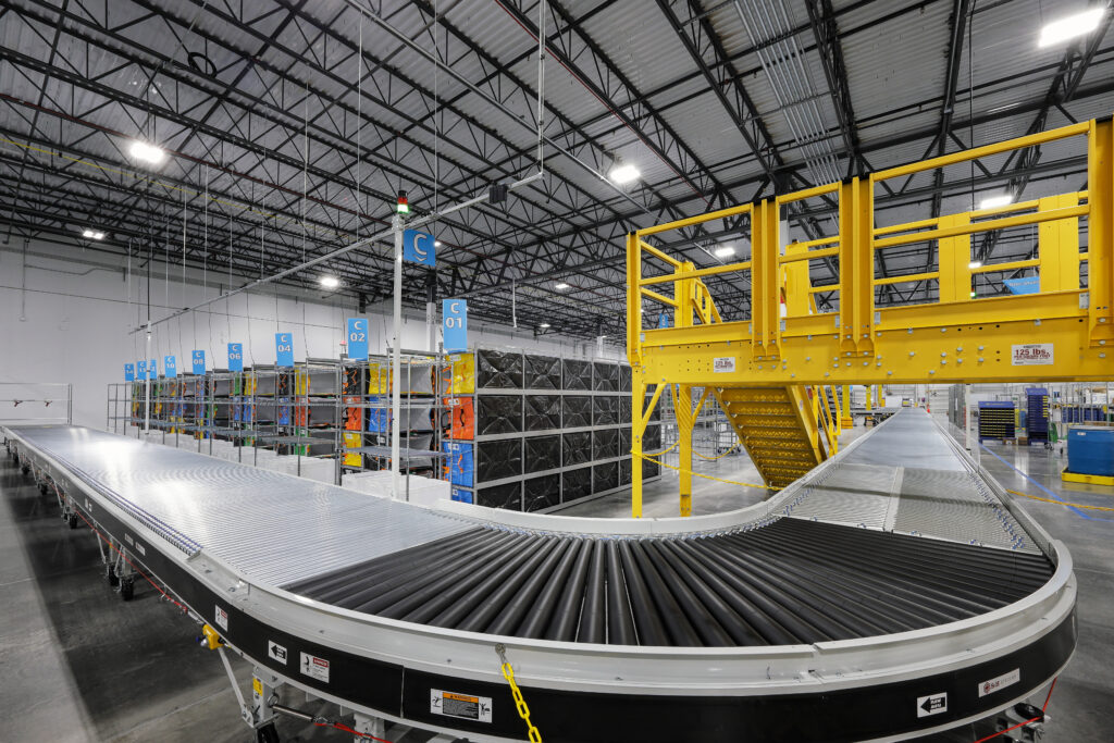 A curved conveyor belt system in a warehouse, with rows of storage shelves and a yellow metal staircase platform.