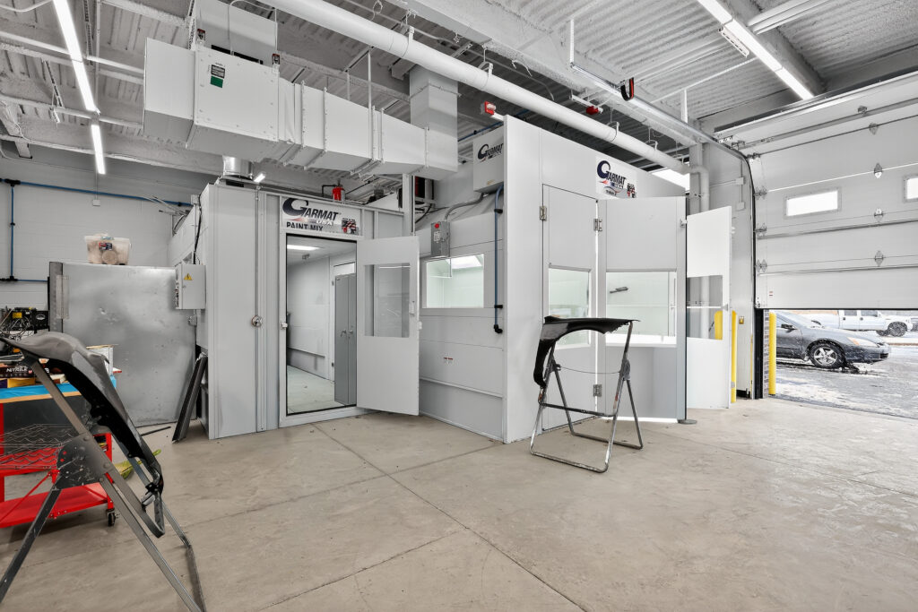 A Garmat paint booth in an automotive workshop, with car bumper parts on stands and an open garage door showing a parking lot.