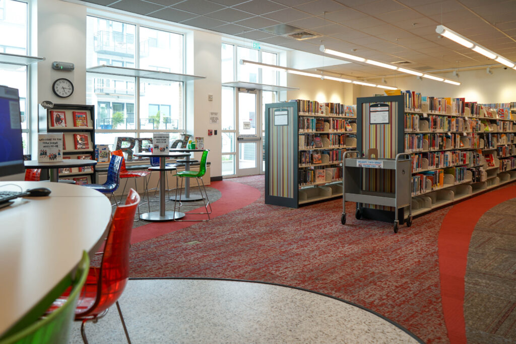 Library interior with rows of bookshelves, a book cart, a cafe-style seating area with colorful plastic chairs, and floor-to-ceiling windows.
