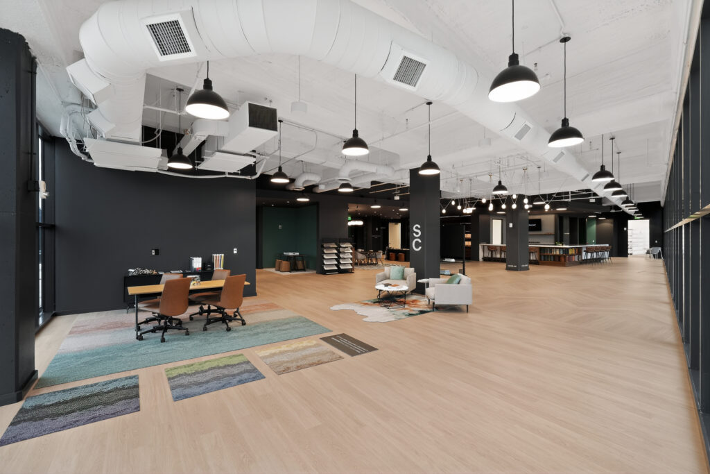 A wide view of a modern, open-concept office showroom with a lounge area, workspace, and flooring samples on the light wood floor.