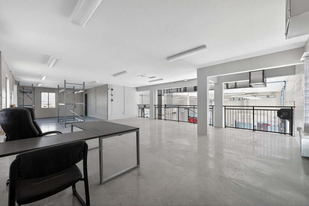 An empty office space with a desk, chairs, and a metal scaffolding frame, overlooking an industrial lower level through a black railing.