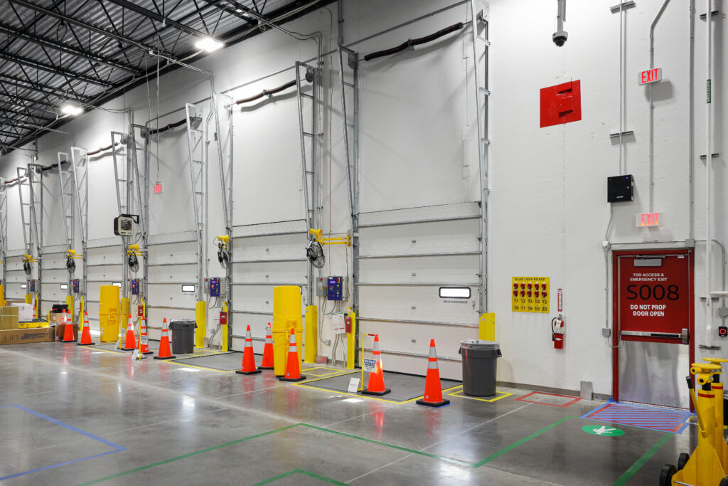 A row of white loading dock doors inside a warehouse, with traffic cones, trash cans, and an emergency exit door marked “S008” on the right.