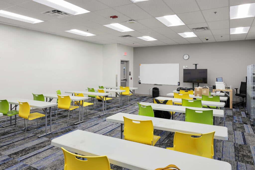Empty classroom with rows of white tables and lime-green chairs, a whiteboard, and a television on a stand.