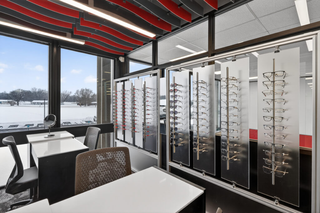 An optometry office interior featuring glass display panels holding rows of eyeglasses and two white desks with chairs, overlooking a snowy landscape.