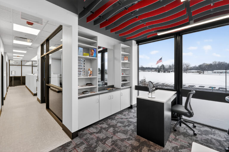 Optical office featuring a display shelving unit with eyeglasses, a desk with a mirror, and a window overlooking a snow-covered parking lot.