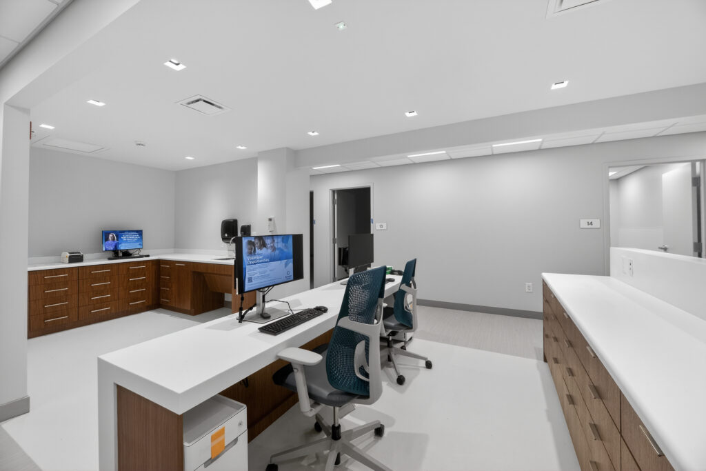 A modern office space with white countertops, wooden cabinetry, and two computer workstations with ergonomic mesh chairs.