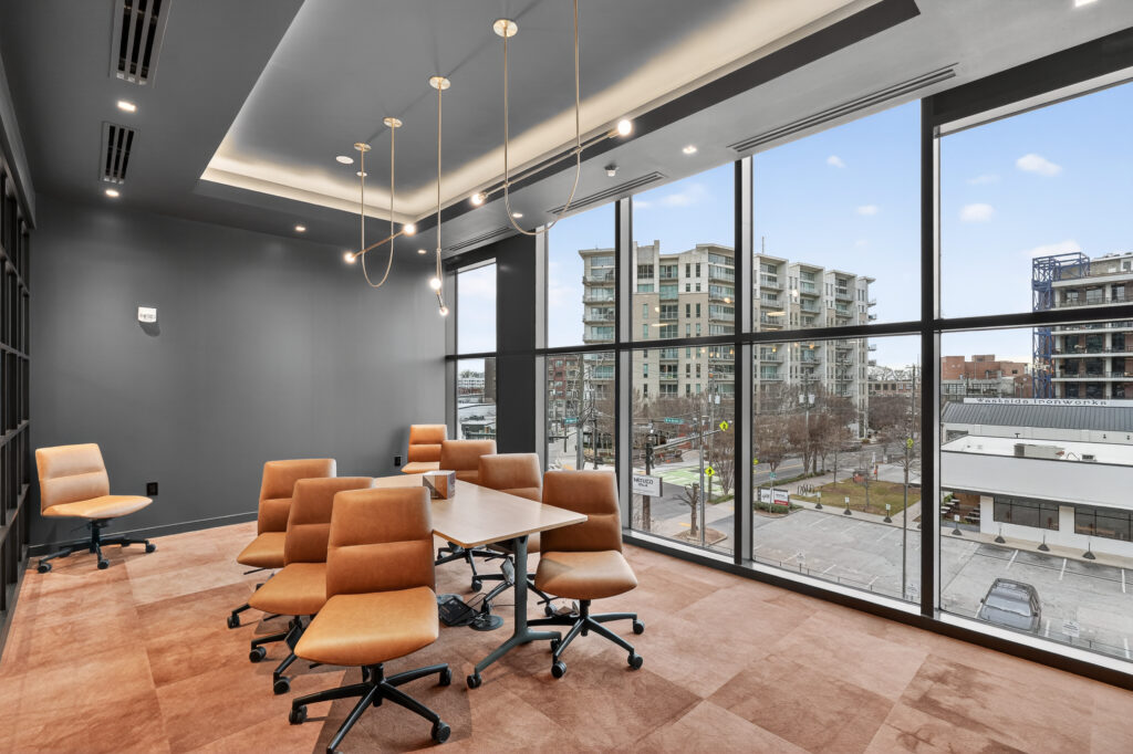 A conference room with a table and tan office chairs, floor-to-ceiling windows looking out over a city street and apartment buildings.