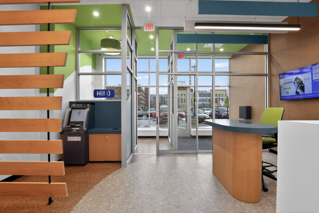 Interior of a bank lobby with an ATM, a reception desk, and a glass-enclosed entrance looking out toward a parking lot.