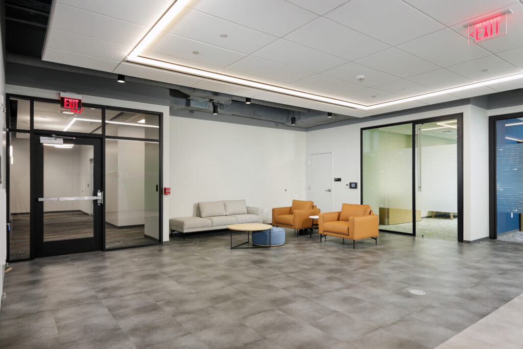Office lobby with a beige sofa, two orange armchairs, a circular coffee table, and a blue ottoman on a gray tiled floor.