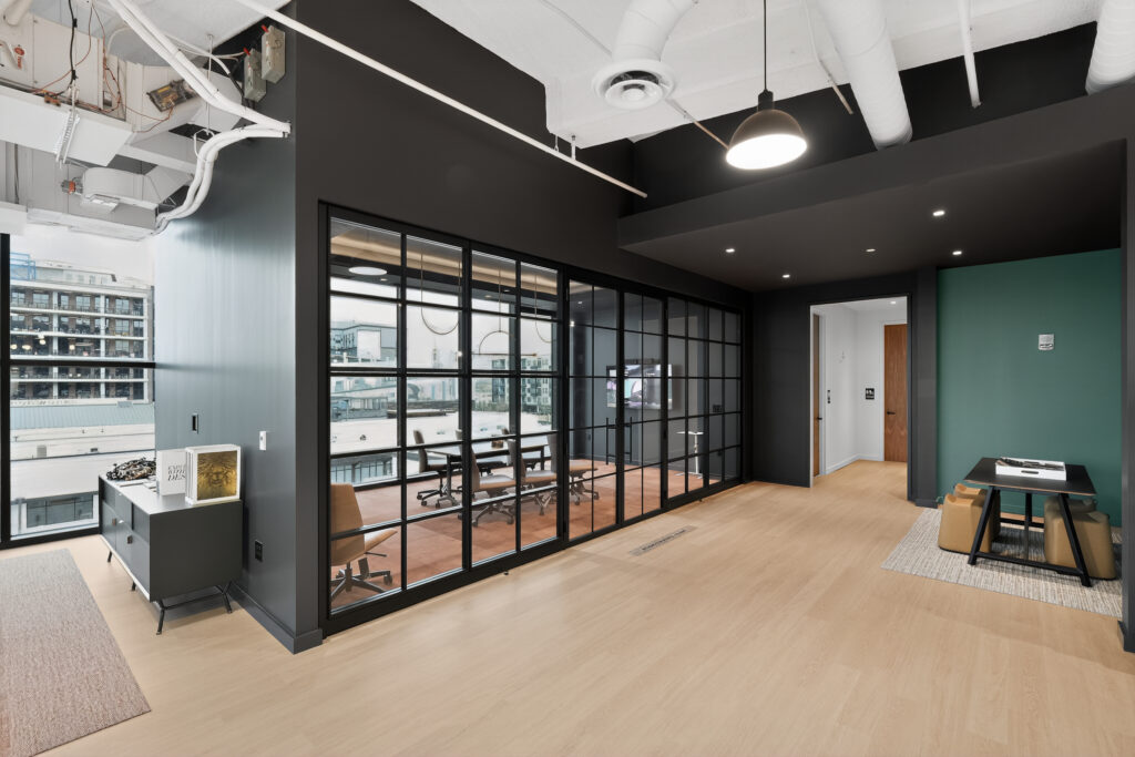 Modern office interior featuring a glass-walled conference room, a small workspace with two stools, and open-plan flooring.
