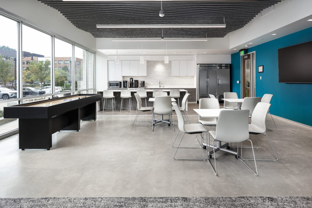 A modern office breakroom with a kitchen area, a shuffleboard table, and several tables with white chairs.