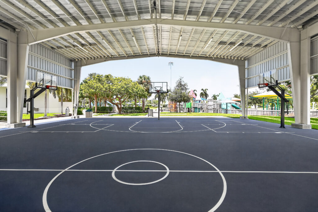 An outdoor basketball court under a large metal roof, featuring two hoops and a dark blue surface.