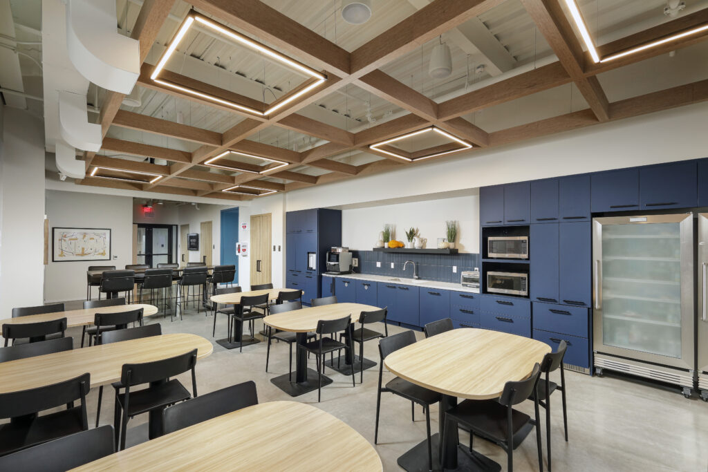 Office breakroom featuring several wooden tables with black chairs, a kitchenette with blue cabinets, and a coffered ceiling with integrated square lighting.