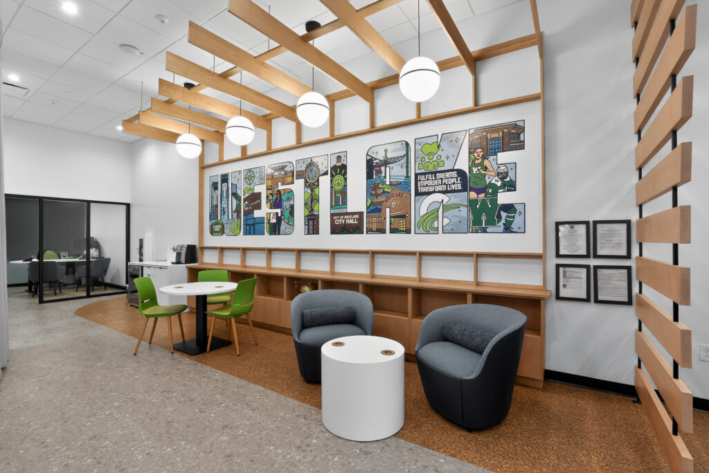 A lounge area with two gray armchairs, a round coffee table, and a bistro table with green chairs, in front of a wall mural that spells “HUSTLE.”