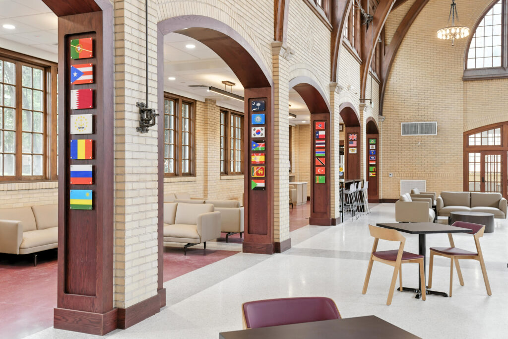 Interior of a large hall with brick walls and arched pillars displaying vertical rows of national flags, featuring seating areas with tables and couches.
