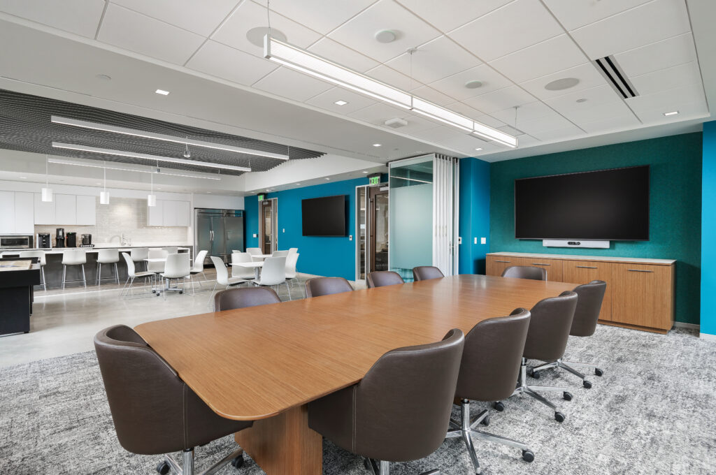 A modern conference room with a long wood-topped table and brown rolling chairs, featuring a teal accent wall with a wall-mounted display and an adjacent kitchenette area with white seating.