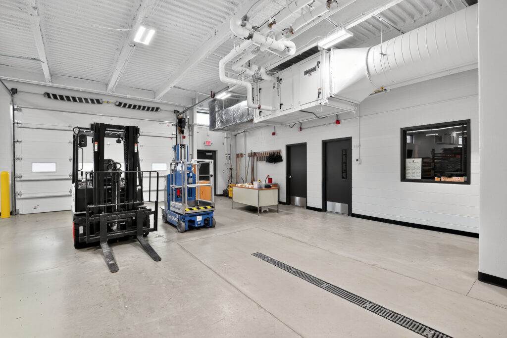 A warehouse interior with a black forklift, a blue scissor lift, a workbench, and two black doors.
