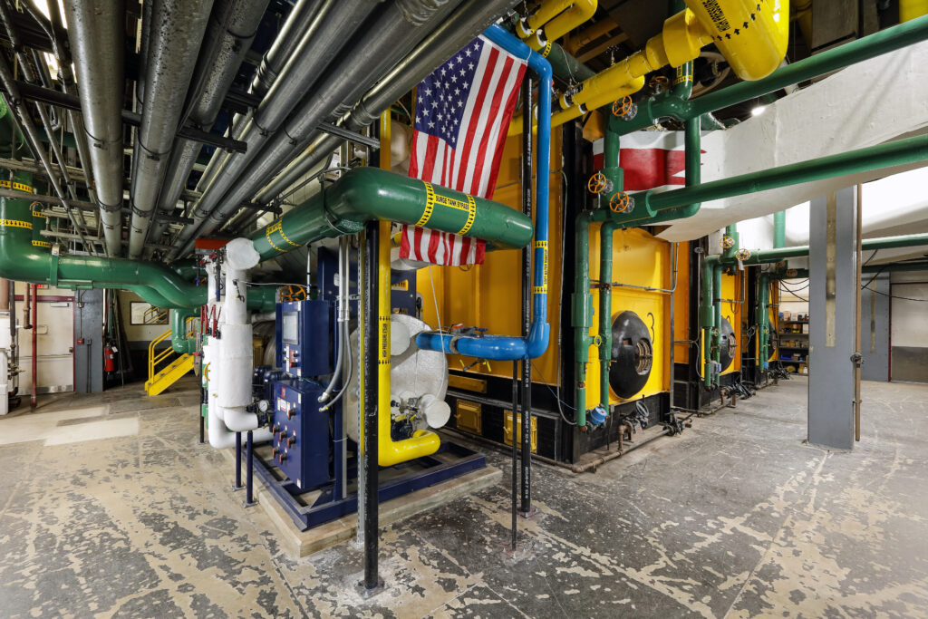 A mechanical room containing large industrial boilers, pipes in various colors, and a hanging American flag.