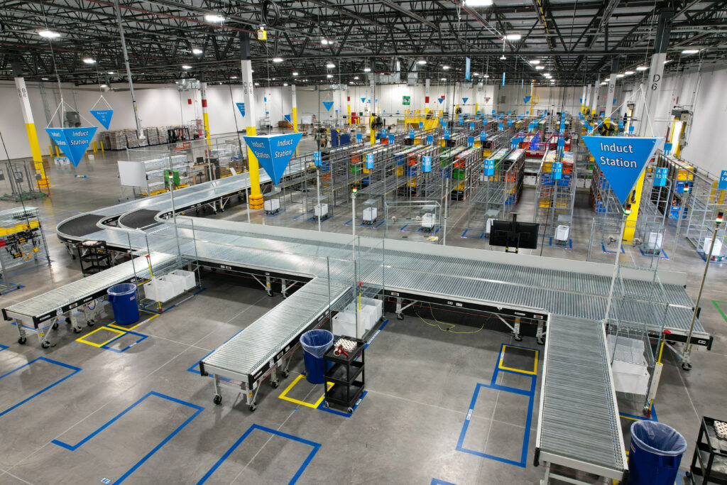 High-angle view of a distribution center with an extensive conveyor belt system, rows of metal storage racks, and overhead blue triangular signs labeled “Induct Station.”