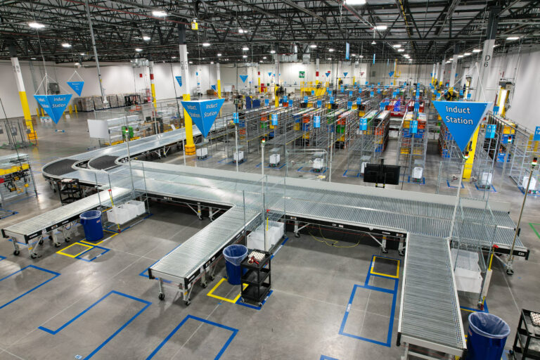 High-angle view of a distribution center with an extensive conveyor belt system, rows of metal storage racks, and overhead blue triangular signs labeled “Induct Station.”