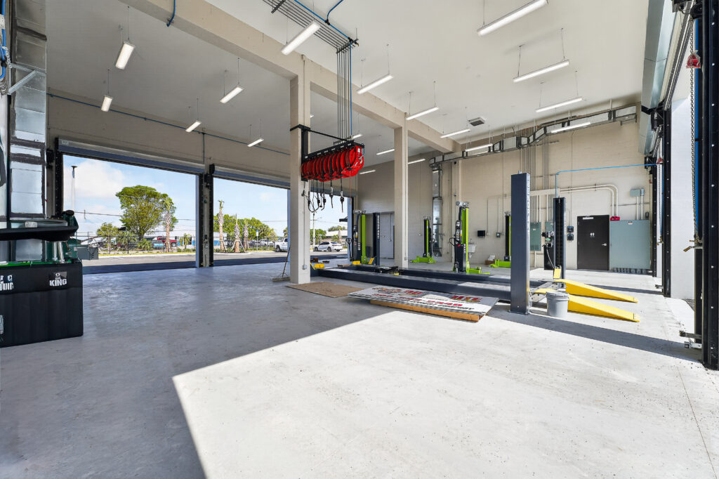 An empty automotive repair shop with multiple hydraulic vehicle lifts, open bay doors, and hanging service reels.