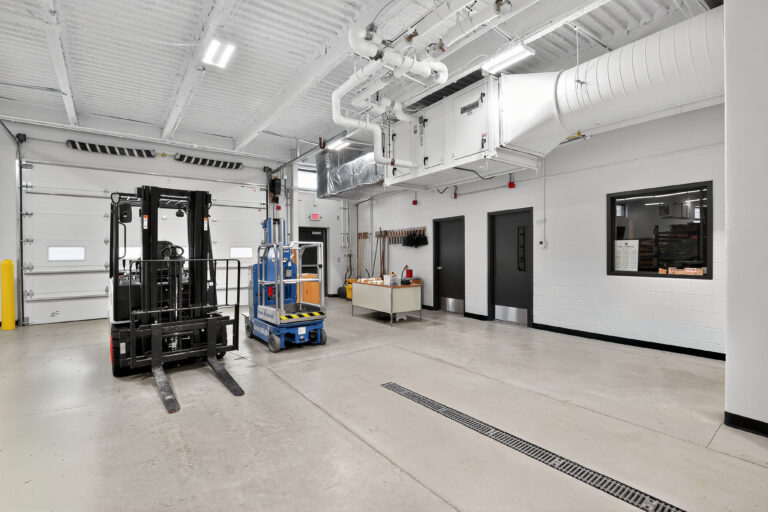 A warehouse interior with a black forklift, a blue scissor lift, a workbench, and two black doors.
