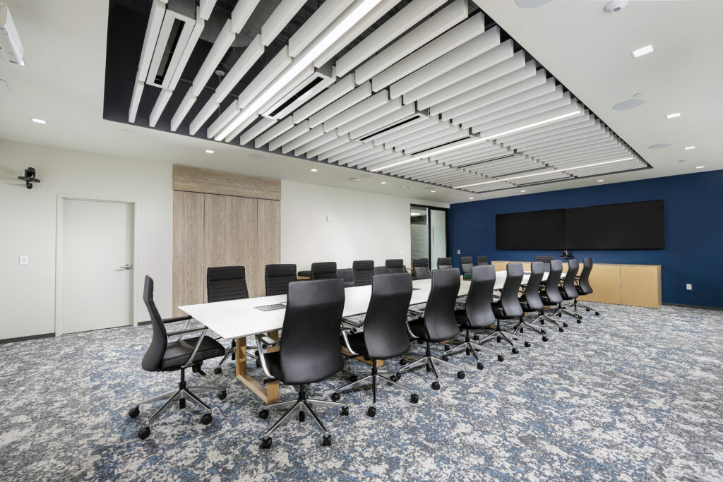 A long conference table with black chairs in an office boardroom with a patterned carpet and a linear ceiling installation.