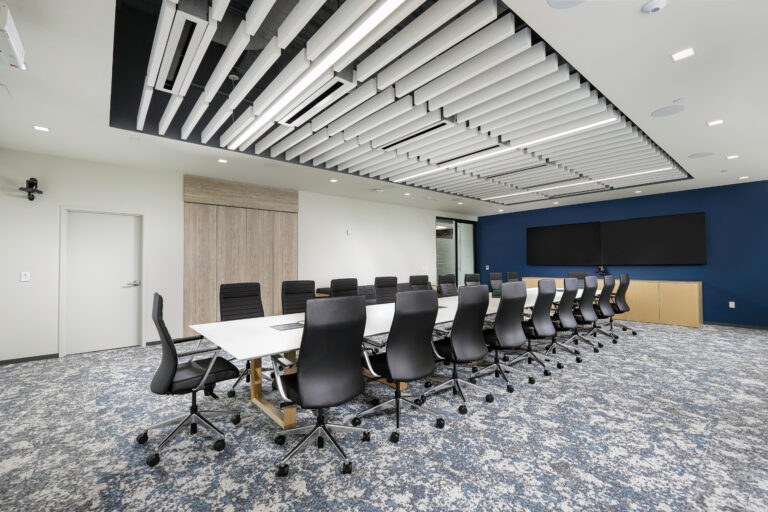 A long conference table with black chairs in an office boardroom with a patterned carpet and a linear ceiling installation.