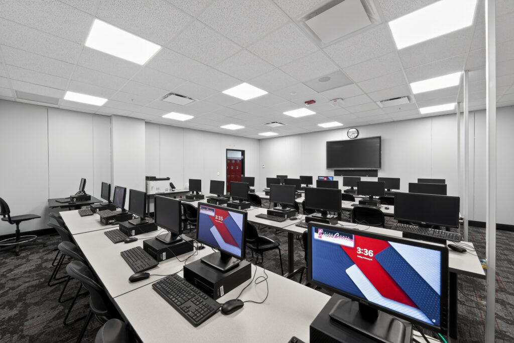 A computer lab filled with rows of desks, each equipped with a monitor, keyboard, and mouse, facing a wall-mounted display.