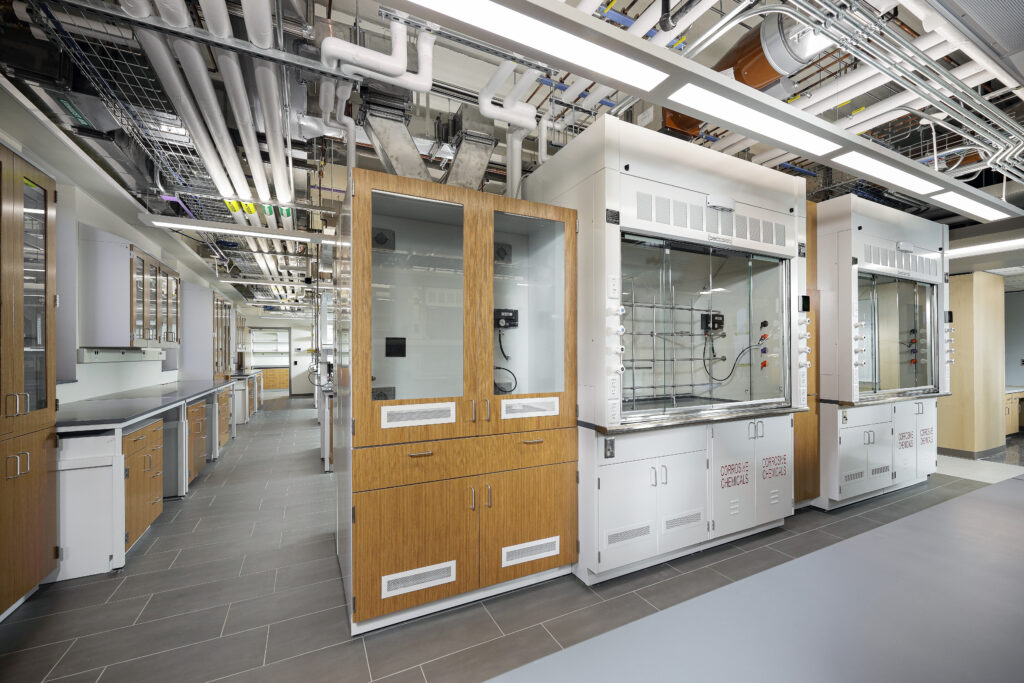 A laboratory interior featuring rows of fume hoods, wooden cabinetry, and exposed ceiling pipes and conduits.