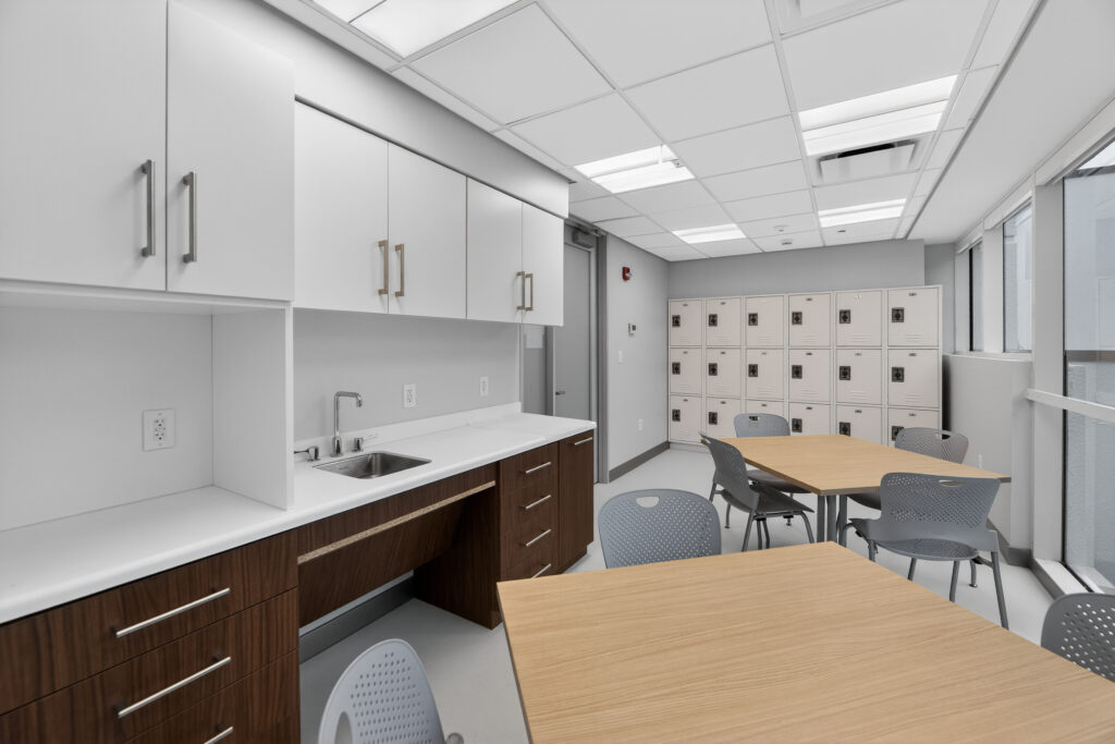 A breakroom with white upper cabinets, a sink, a row of storage lockers, and light wood tables with gray chairs.