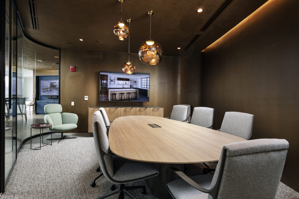 A modern conference room with a wooden oval table, six grey office chairs, a television on a dark wall, and a light green accent chair in the corner.