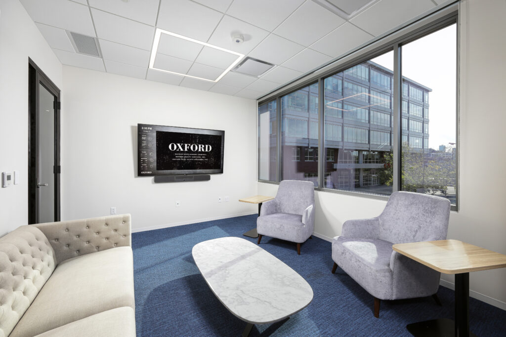 Office lounge with a tufted sofa, two armchairs, a marble coffee table, and a wall-mounted screen displaying the Oxford company logo.