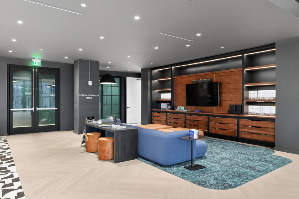 Modern lounge area with a blue sectional sofa, two round tan stools, a desk, and a television mounted on a wood-paneled wall.